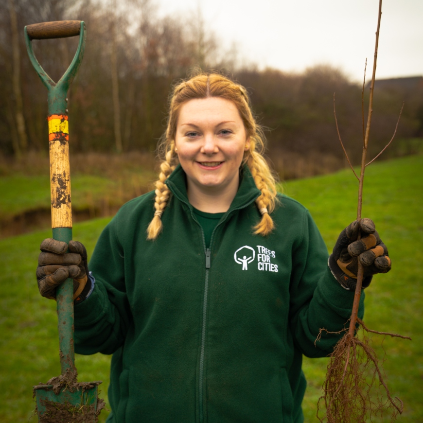 A young lady with pigtails smiles at the camera. She's holding a tree sapling and a shovel, and looks ready to get planting with gloves and a 'Trees for Cities' fleece.