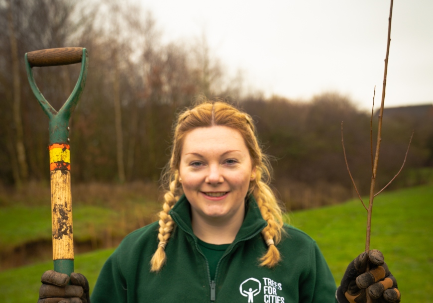 A young lady with pigtails smiles at the camera. She's holding a tree sapling and a shovel, and looks ready to get planting with gloves and a 'Trees for Cities' fleece.