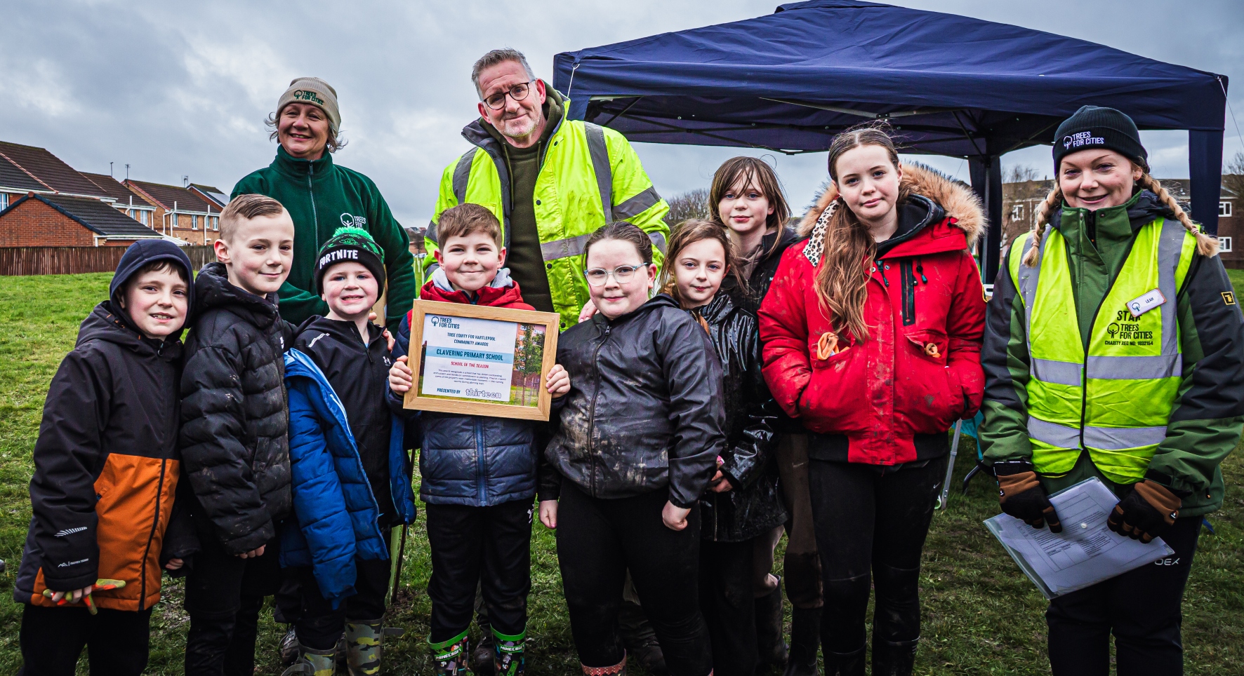 A group of young people pose for a photo with group leaders. A boy in the middle proudly holds a certificate. They're wrapped up for a cold, blustery day but look cheerful.