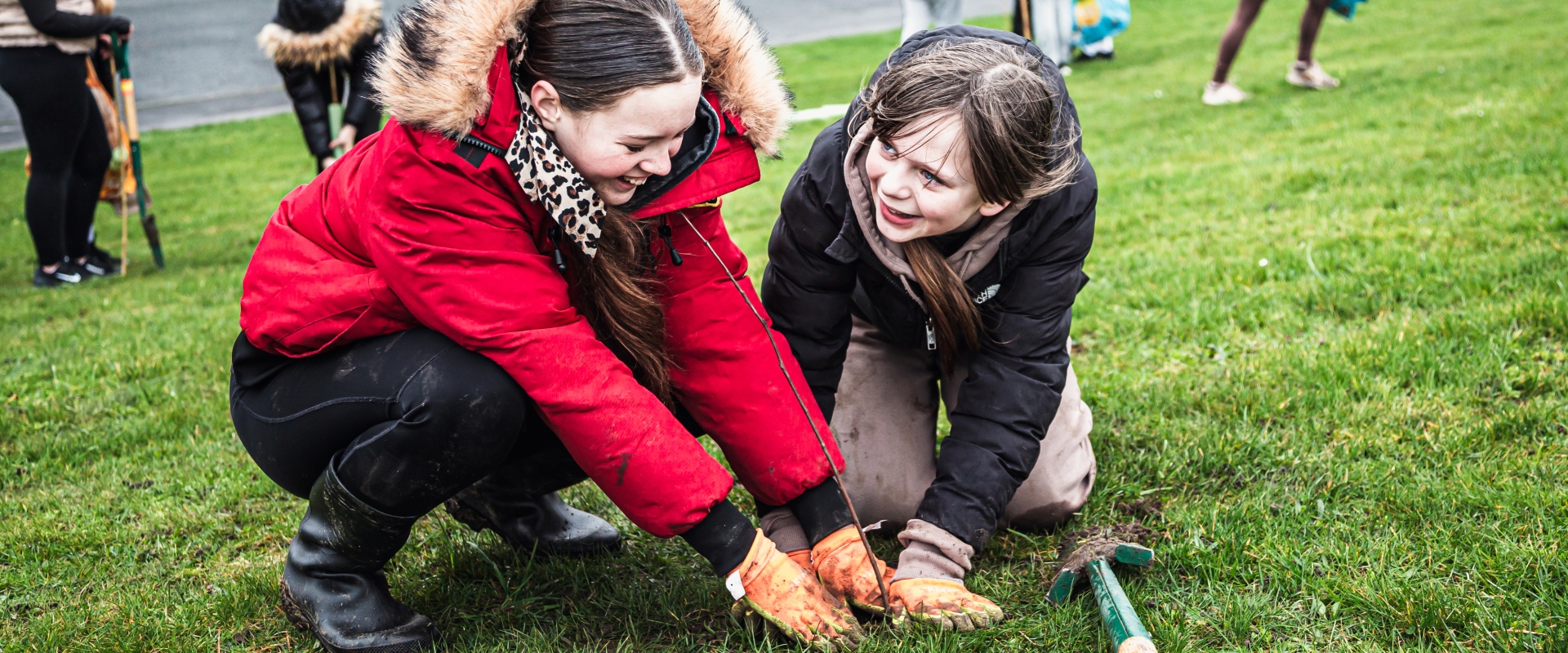Close-up of two children as they pat the ground around a newly-planted tree sapling. One wears a bright red coat and an even brighter smile. The other also looks happy with their work.