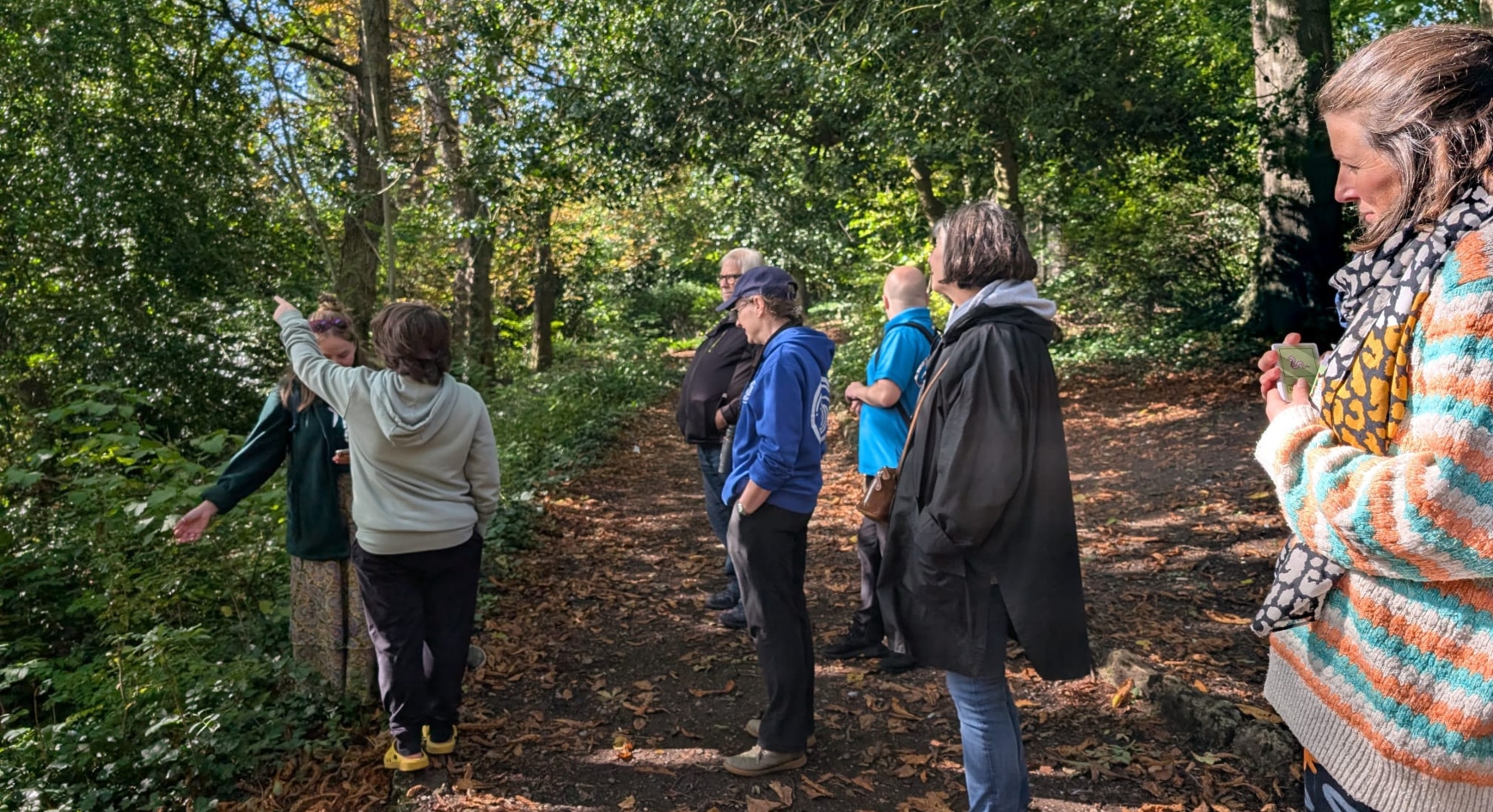 A group of people stand in a woodland, looking in the direction that a young person in a grey hoodie is pointing.
