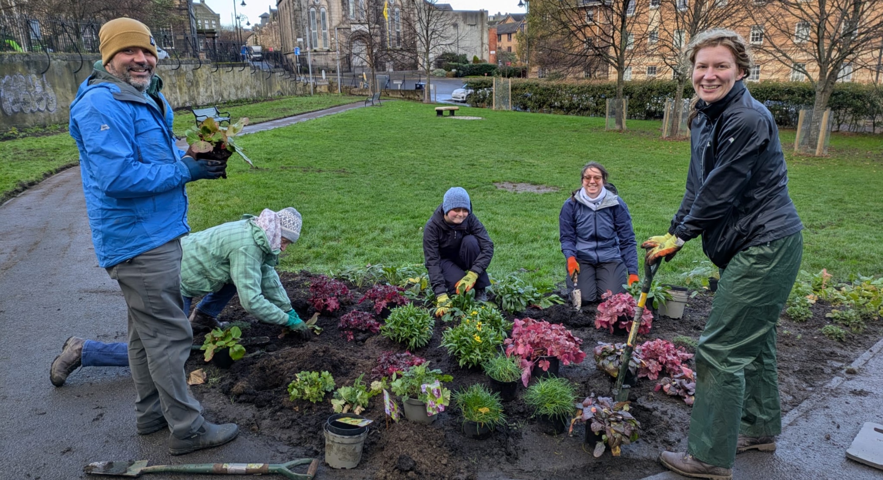 A small group tends to a corner flower bed in small green space. They're in an urban area and it looks chilly, but there are wide smiles all round.