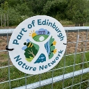Close-up of a white circular 'Part of Edinburgh Nature Network' sign, installed on the metal gate of a woody field