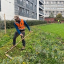 A man in orange high-vis scythes grasses in a large green space in the middle of what looks like residential flats.
