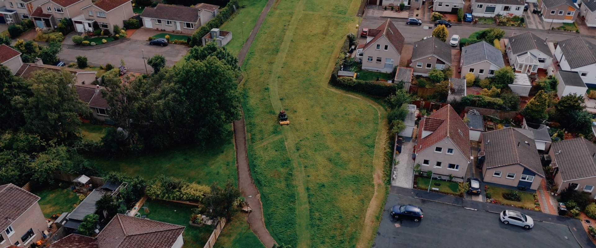 An aerial view of a grassy field surrounded by houses and residential roads. A lone mower cuts a track down the middle.