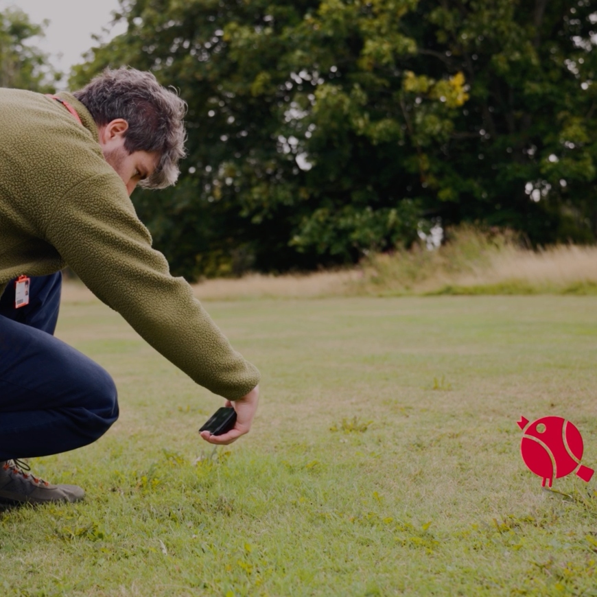 A man crouches down in a grassy field, with trees behind. He looks to be measuring the quality of the grass. A red icon of a robin watches on.
